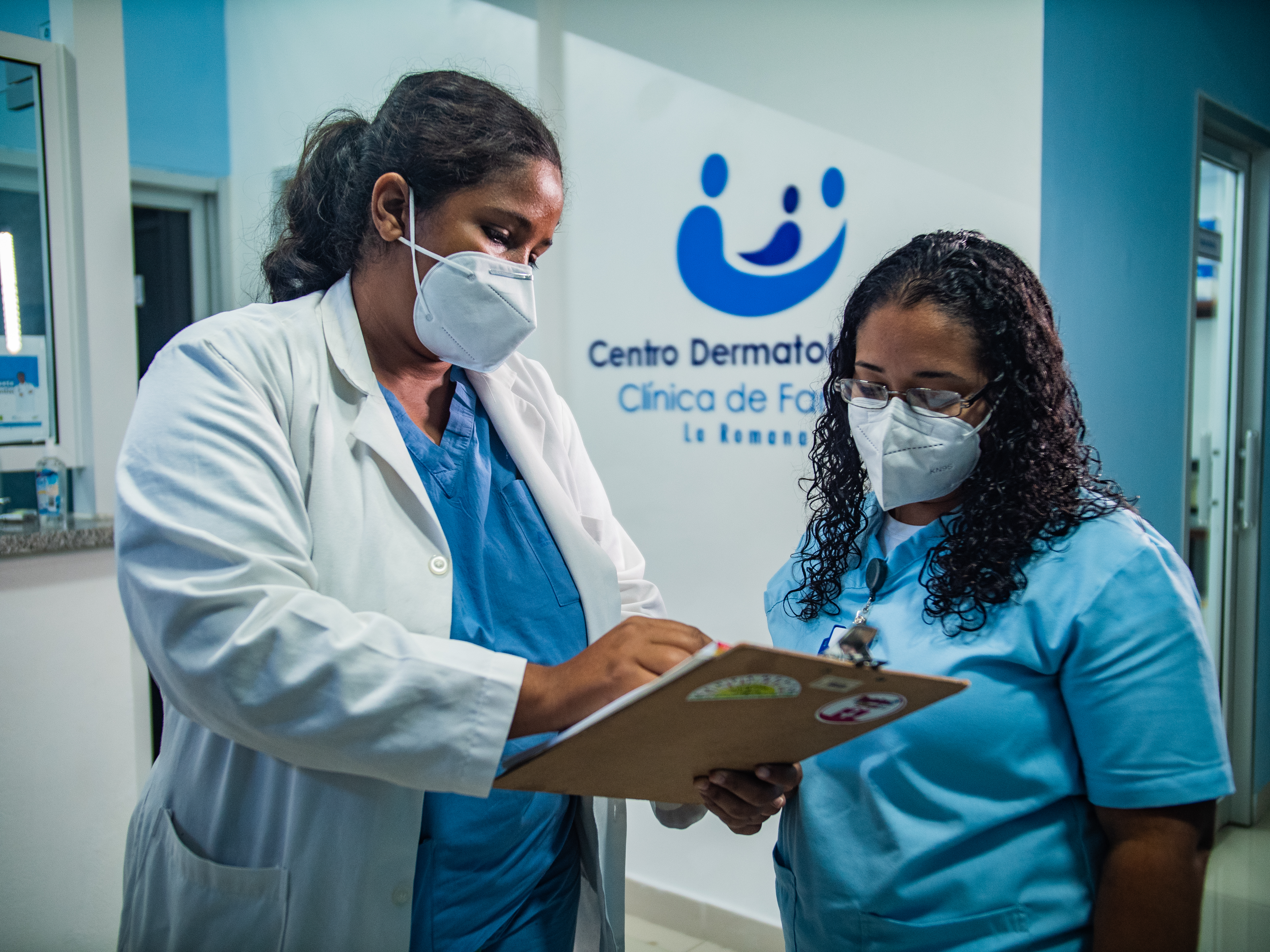 Two female health providers at Clínica de Familia look at a clipboard together. One wears a lab coat and the other does not. They wear masks.