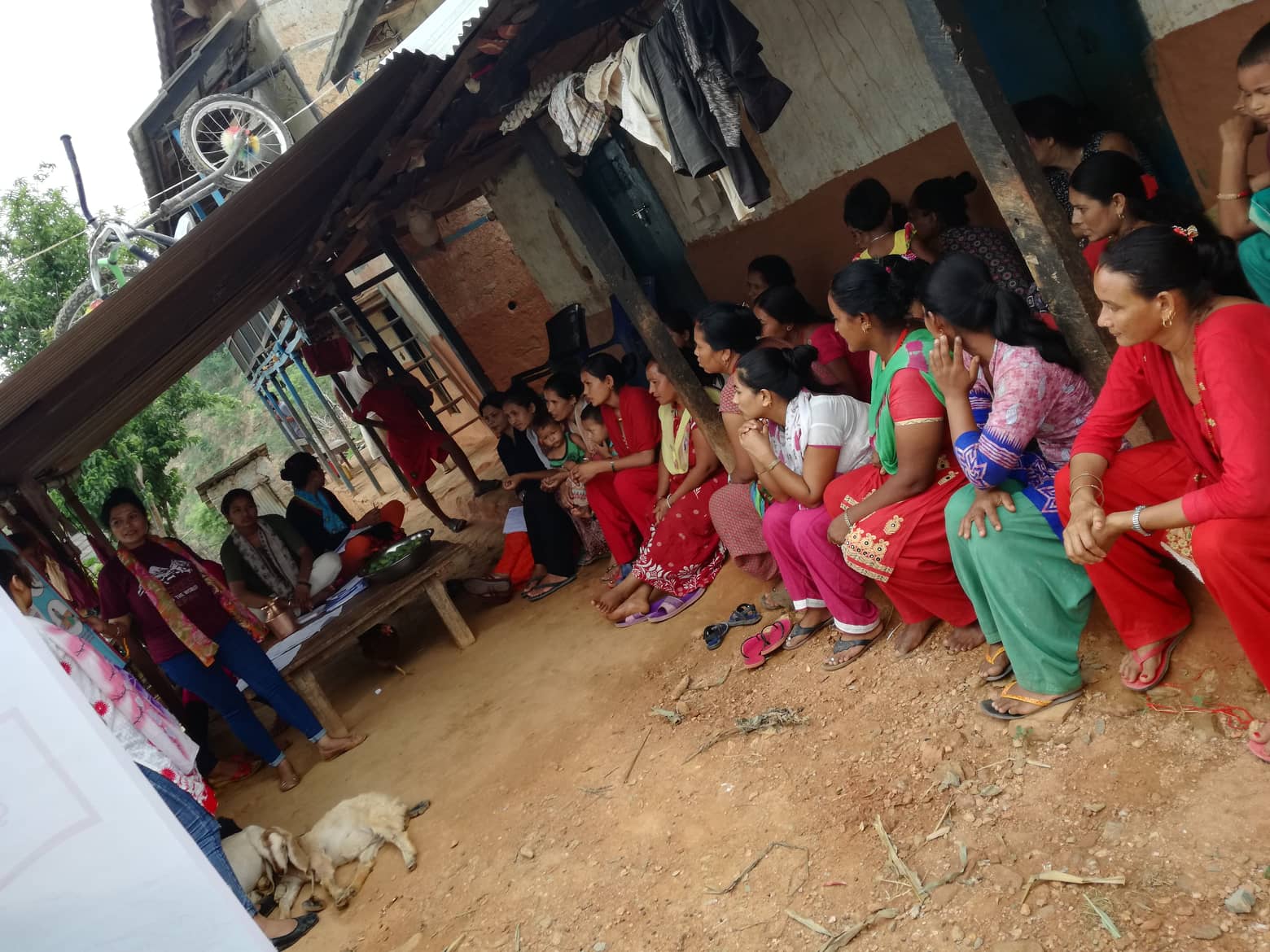 Women in Ramechaap District sitting on a step outside a building listening to someone talking. 