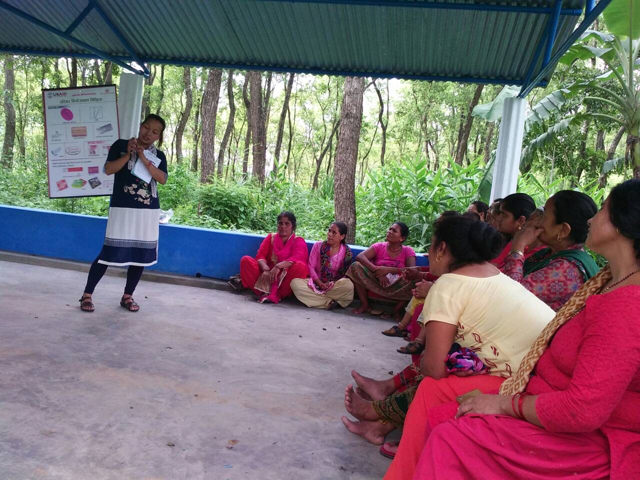 Community change agent demonstrating FP material to women sitting on the floor in a cement structure with a roof.