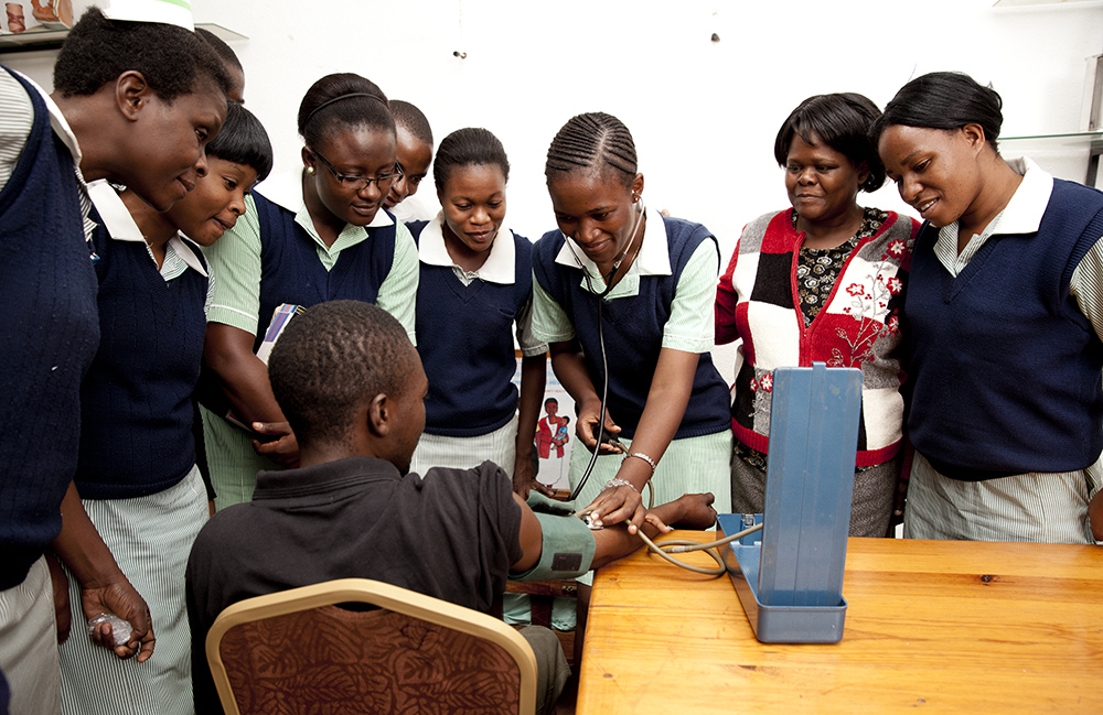 Photo of female students standing and watching a student that is taking the blood pressure of a young man sitting in a chair.
