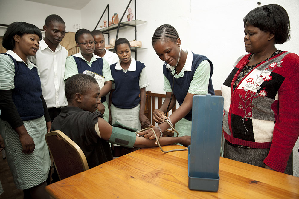 Nurses learning how to take blood pressure of a man sitting in a chair.